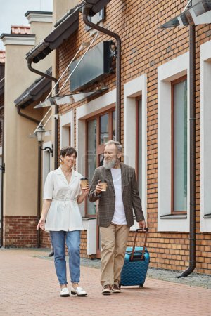 älteres Paar, glücklicher Mann und Frau zu Fuß mit Kaffee to go und Gepäck auf der Straße, urbaner Lebensstil