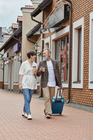 happy elderly couple, man and woman walking with coffee to go and luggage on street, urban lifestyle