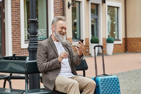 glücklicher älterer Mann mit Smartphone und auf Bank in der Nähe von Gepäck, mit Gadget, Alter in der Technik