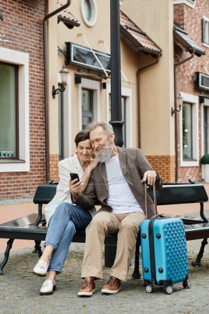 pareja mayor, hombre barbudo feliz celebración de teléfono inteligente, sentado con la mujer en el banco, equipaje, gadget