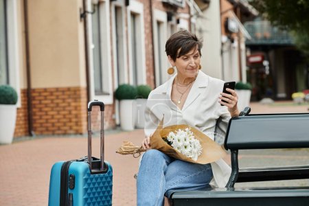 elderly woman with short hair using smartphone, holding bouquet, sitting on bench near luggage
