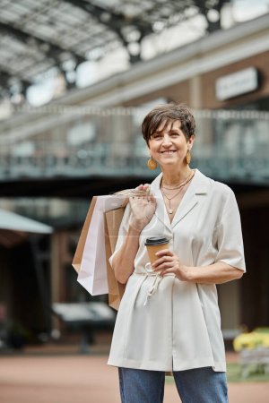 Téléchargez les photos : Heureuse femme âgée avec les cheveux courts tenant des sacs à provisions et du café pour aller près de la sortie à l'extérieur - en image libre de droit