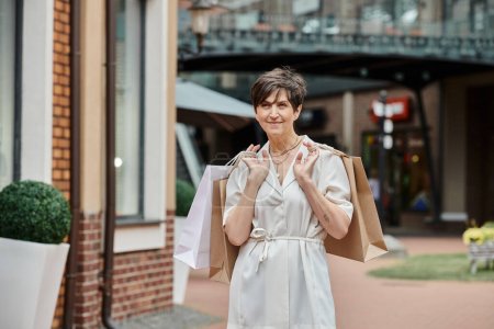 aging population, pleased senior woman holding shopping bags near outlet, active senior, positive