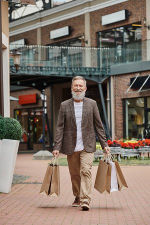 glücklicher bärtiger Mann, der mit Einkaufstaschen spaziert, Seniorenleben, urbane Straße, positives, stylisches Outfit
