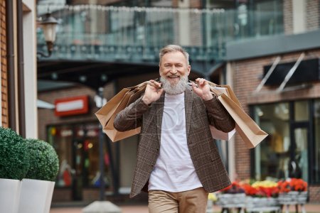 homme positif et barbu marchant avec des sacs à provisions, la vie des personnes âgées, rue urbaine, tenue élégante