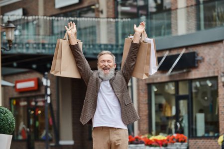 Téléchargez les photos : Homme excité et barbu marchant avec des sacs à provisions, la vie des personnes âgées, rue urbaine, tenue élégante - en image libre de droit