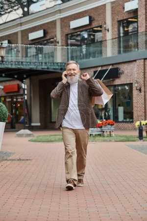 homme heureux et âgé avec barbe parler sur smartphone, tenant des sacs à provisions, marchant près de la sortie