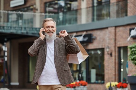 homme âgé positif avec barbe parlant sur smartphone, tenant des sacs à provisions, marchant près de la sortie