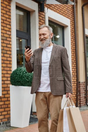 homme âgé gai avec barbe en utilisant un smartphone, tenant des sacs à provisions, marchant près de la sortie