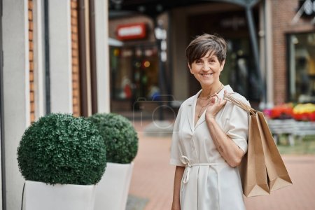 glückliche Seniorin mit kurzen Haaren, die Einkaufstaschen in der Hand hält und in die Kamera schaut, Outdoor-Einkaufszentrum, Outlet