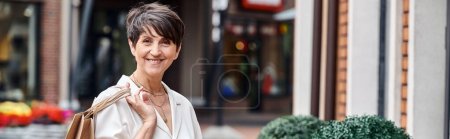 happy senior woman with short hair holding shopping bags and looking at camera, outdoor mall, banner