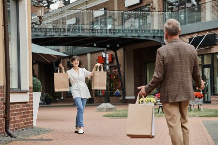 happy elderly woman showing shopping bags to man, husband and wife in outlet, outdoors, lifestyle