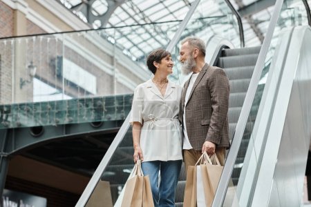 hombre y mujer senior positivo de pie en escaleras mecánicas, bolsas de compras, mirándose en el centro comercial