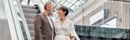 happy senior man and woman standing on escalator, looking at each other in mall, banner, horizontal