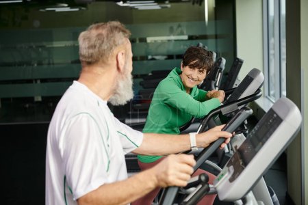 fröhliche Frau blickt auf ältere Männer, Mann und Frau, die im Fitnessstudio trainieren, aktive Senioren, Sport