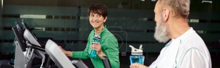 happy woman looking at elderly man, husband and wife in gym, holding sports bottles, banner