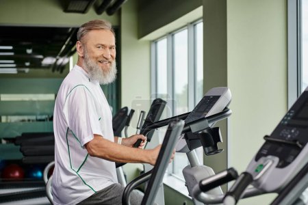 Téléchargez les photos : Homme barbu heureux travaillant sur la machine d'exercice, les personnes âgées dans la salle de gym, senior actif, fitness et sport - en image libre de droit