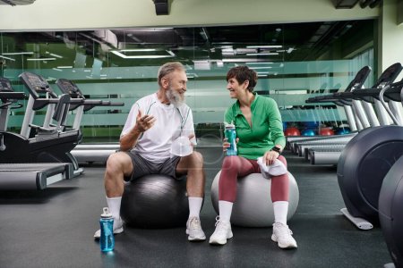 couple âgé sportif, homme et femme joyeux assis sur des balles de fitness, tenant des bouteilles avec de l'eau