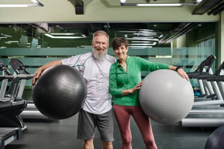 sporty elderly couple, cheerful man and woman holding fitness balls, active seniors in gym