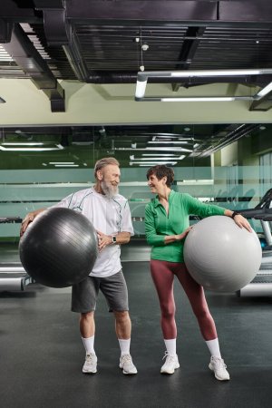 pareja de ancianos, hombre alegre y mujer sosteniendo pelotas de fitness, personas mayores activas mirándose