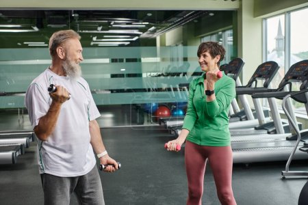 Foto de Pareja de ancianos, hombre y mujer alegres haciendo ejercicio con pesas, personas mayores activas mirándose - Imagen libre de derechos