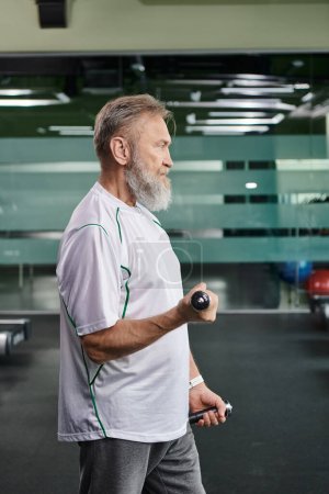 Foto de Vista lateral del anciano barbudo haciendo ejercicio con pesas en el gimnasio, senior activo, rutina de fitness - Imagen libre de derechos