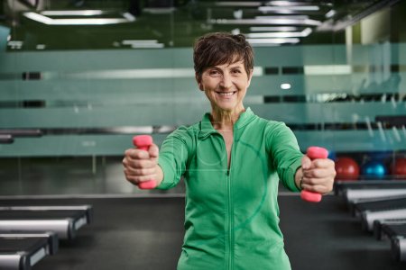 mujer feliz con el pelo corto haciendo ejercicio con pesas, mirando a la cámara en el gimnasio, retrato