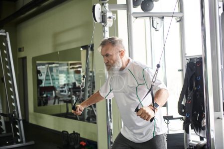 motivierter älterer Mann mit Bart, der im Fitnessstudio auf dem Trainingsgerät trainiert, Sportler, aktiver Senior