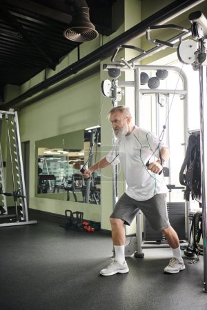 concentrated elderly man with beard working out on exercise machine in gym, athlete, motivation