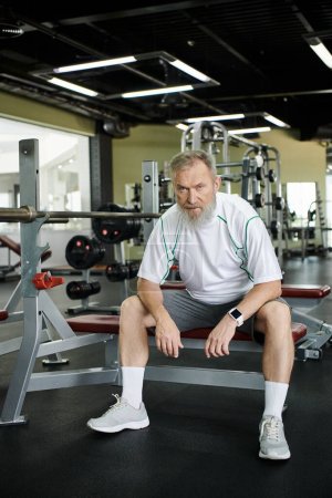 tired elderly man with beard looking at camera after workout, sitting on exercise machine in gym