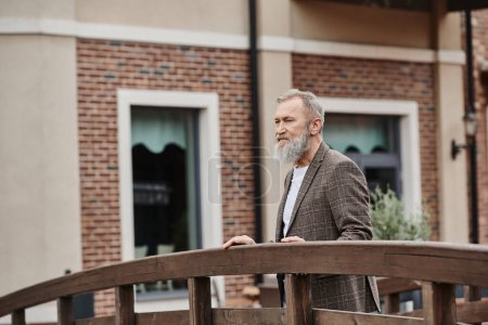 bearded elderly man with grey hair standing on wooden bridge, looking away, thinking, urban backdrop