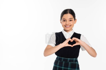 happy schoolgirl in black vest showing heart sign with hands and looking at camera isolated on white
