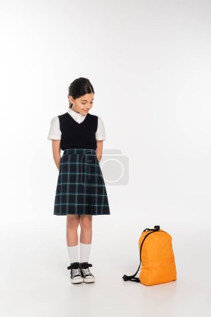 glückliches Mädchen in Schuluniform, stehend und mit Blick auf den Rucksack auf weißem Hintergrund, zurück zur Schule