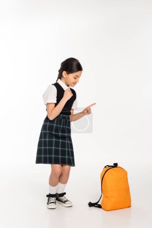 glückliches Mädchen in Schuluniform, das auf weißem Hintergrund steht und auf den Rucksack blickt und wegzeigt