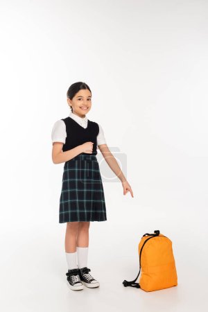 fröhliches Mädchen in Schuluniform steht und zeigt auf Rucksack auf weißem Hintergrund, volle Länge