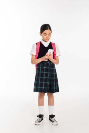 Photo for Digital age, schoolgirl with backpack holding smartphone on white, student in uniform, full length - Royalty Free Image