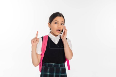 digitales Zeitalter, staunendes Schulmädchen mit Rucksack spricht auf Smartphone isoliert auf weiß und zeigt nach oben