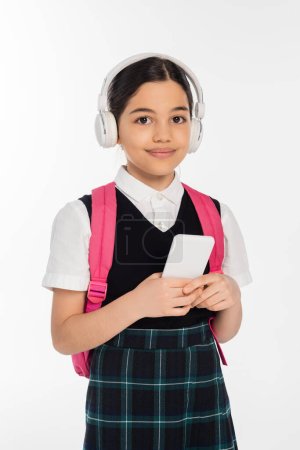 digital age, joyful schoolgirl in wireless headphones holding smartphone isolated on white, student