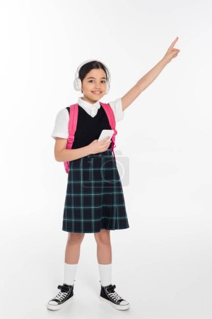 digital age, cheerful schoolgirl in wireless headphones holding smartphone on white, pointing away