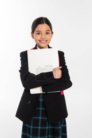 alegre colegiala sosteniendo portátil y mirando a la cámara, chica en uniforme escolar, aislado en blanco
