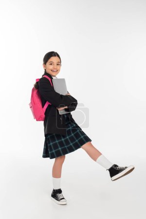 joyful schoolgirl holding laptop and looking at camera, girl in school uniform on white background