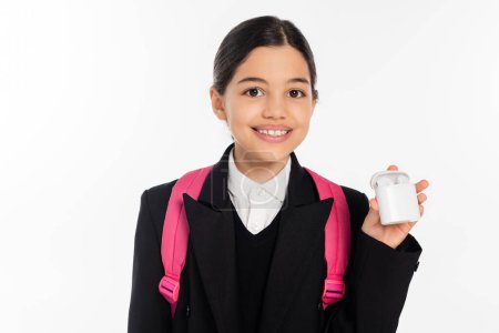 cheerful schoolgirl in uniform holding case with wireless earphones isolated on white, positive