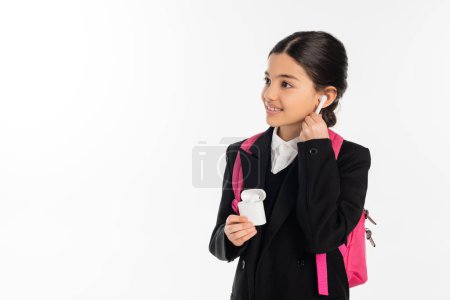 smiling schoolgirl in uniform holding case with wireless earphones isolated on white, positive