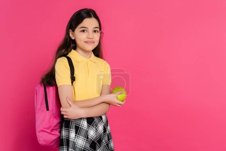 happy schoolgirl with backpack holding green fresh apple isolated on pink, vibrant backdrop