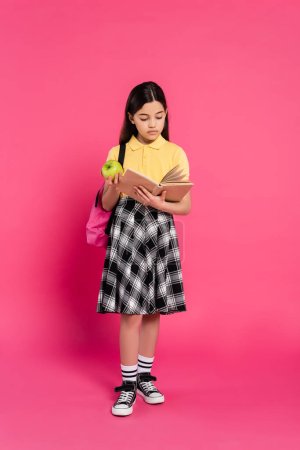 full length, brunette schoolgirl holding green apple and reading book on pink background, backpack