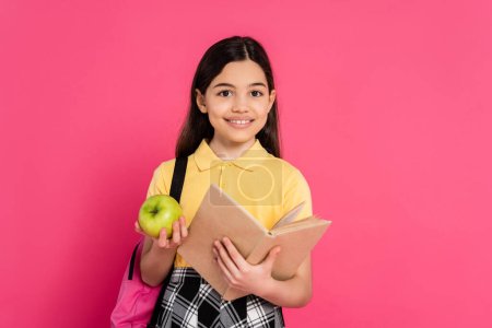 Freude, brünettes Schulmädchen mit grünem Apfel und Lesebuch isoliert auf rosa, Mädchen mit Rucksack