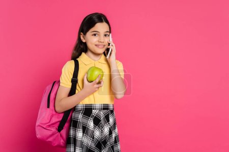 Téléchargez les photos : Heureuse écolière debout avec sac à dos, tenant pomme et parler sur smartphone, appel téléphonique - en image libre de droit