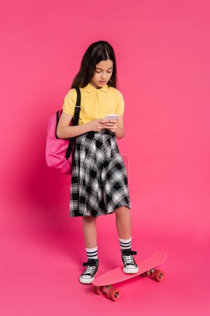 brunette schoolgirl using smartphone and standing near penny board on pink background, youthful life