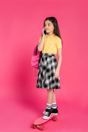 brunette schoolgirl talking on smartphone and riding penny board on pink background, phone call