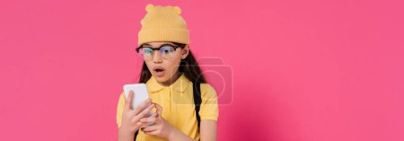 shocked schoolgirl in beanie hat and glasses using smartphone on pink background, stylish, banner